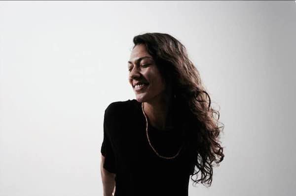 Mohawk water protector, land defender, and rapper Dioganhdih, a dark-skinned two-spirit individual with long brown hair and a thin rope-like necklace who is smiling with their eyes closed